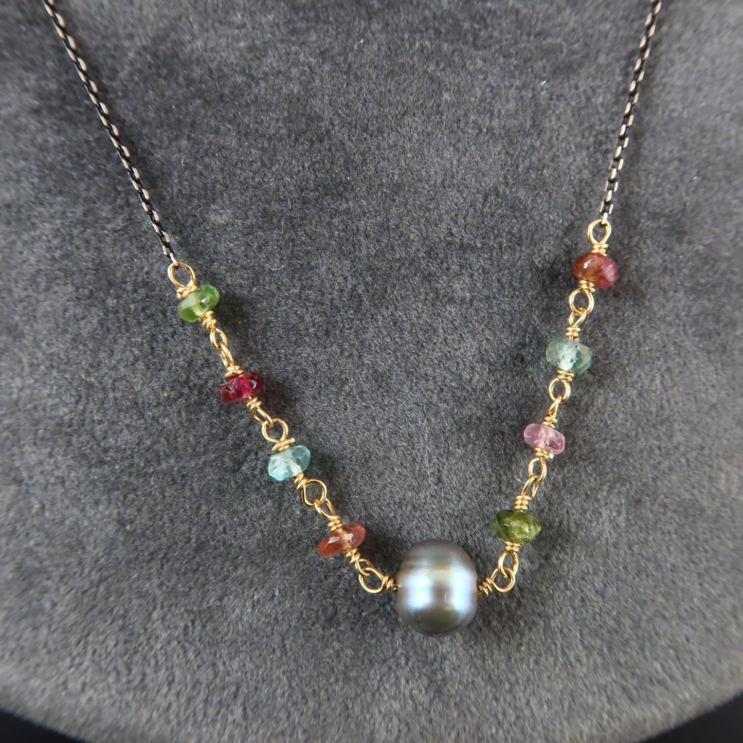 A delicate necklace with a dark chain, featuring a central round black pearl bead and multiple small, colorful wire-wrapped gemstone beads in green, red, blue, and pink, displayed on a textured gray background.