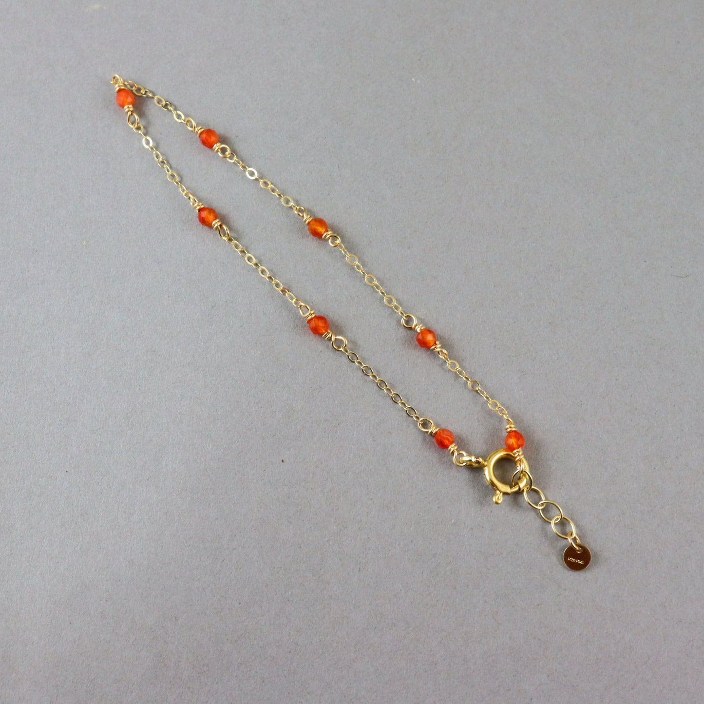 A delicate gold chain necklace with small orange beads spaced along the chain, featuring a gold lobster clasp and a small dangling charm, displayed on a neutral gray background.