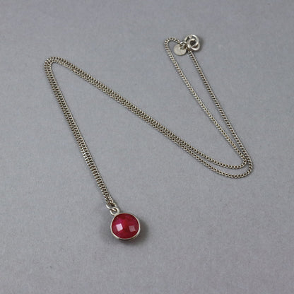 A silver-toned chain necklace with a round faceted red gemstone pendant, displayed on a neutral gray background.