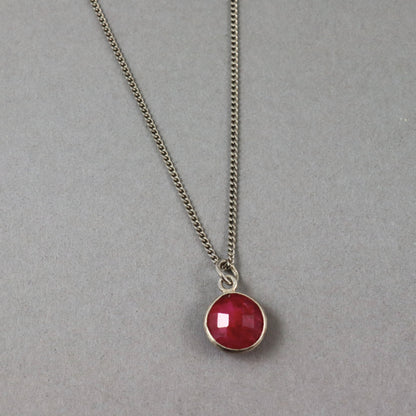 A silver-toned chain necklace with a round bezel-set red gemstone pendant, displayed on a neutral gray background.