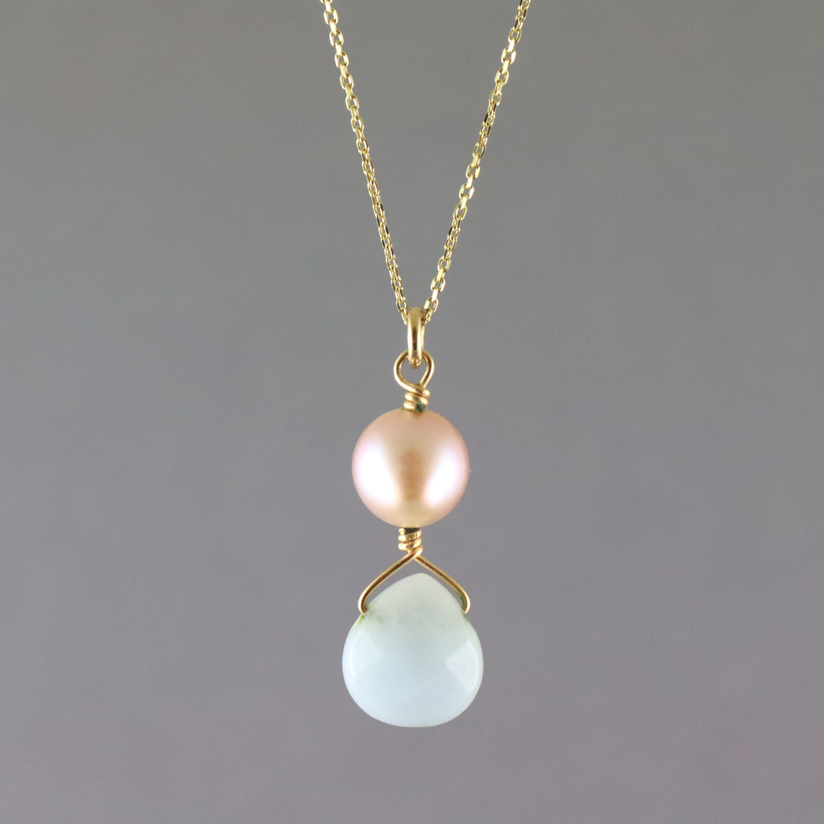 A delicate gold chain necklace featuring a dual pendant design: a round, lustrous pale pink pearl on the top and a faceted, teardrop-shaped light blue stone on the bottom, both set in gold.