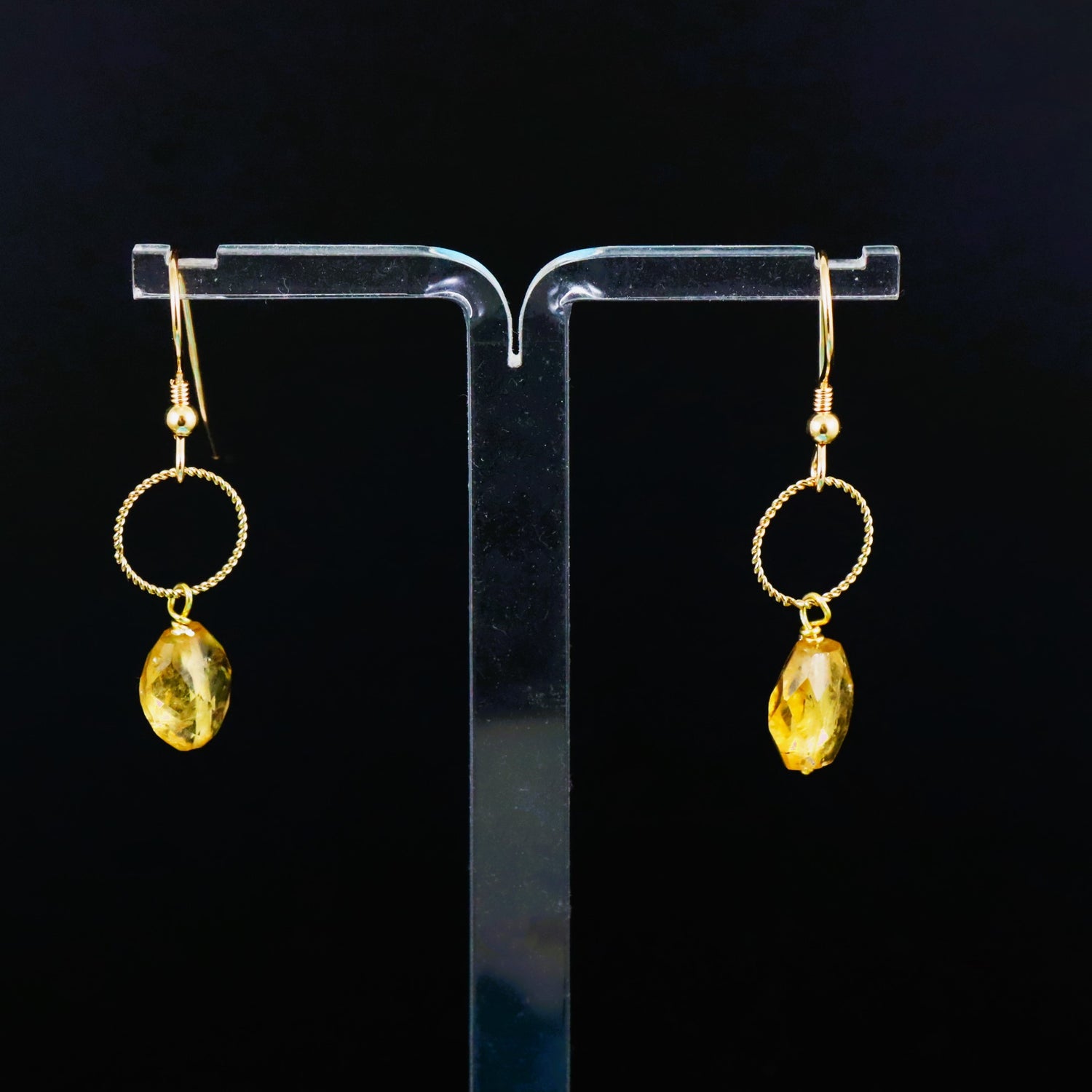A pair of gold-filled twist ring detail citrine gemstone earrings displayed on a clear stand against a black background.
