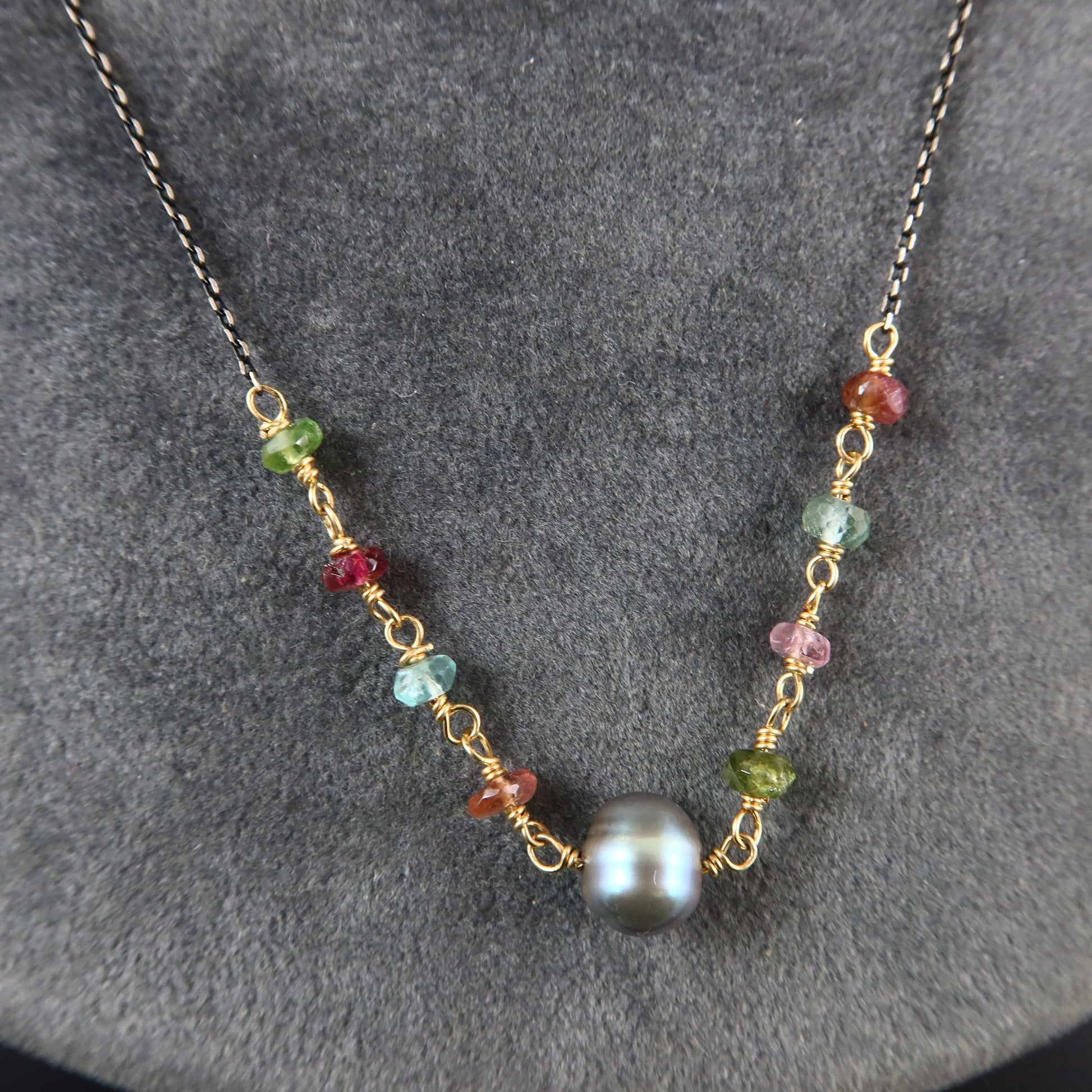 A delicate necklace with a dark chain, featuring a central round black pearl bead and multiple small, colorful wire-wrapped gemstone beads in green, red, blue, and pink, displayed on a textured gray background.