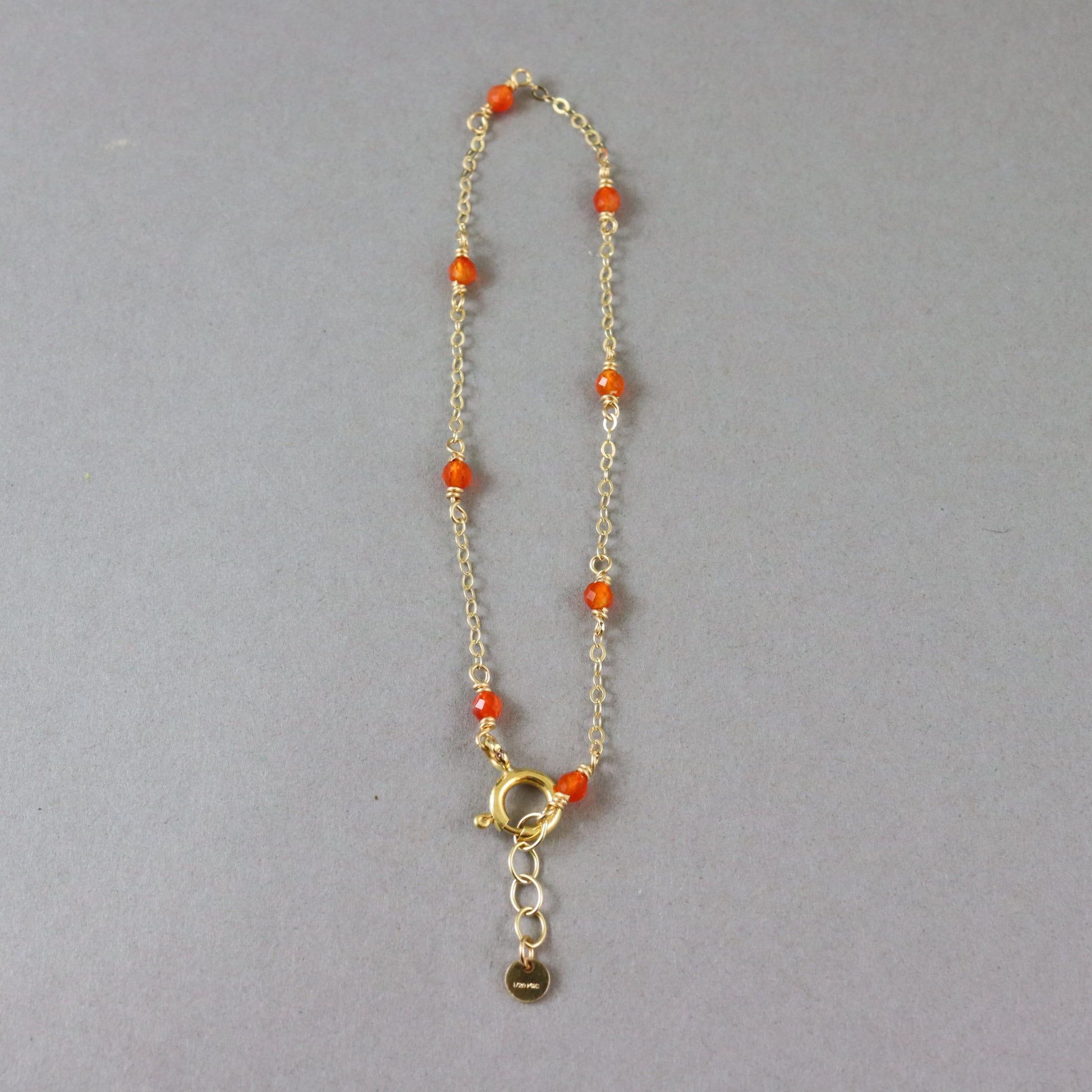 A delicate gold chain bracelet with small orange beads spaced along the chain and a gold clasp with an extender and small hanging tag, shown on a plain gray background.