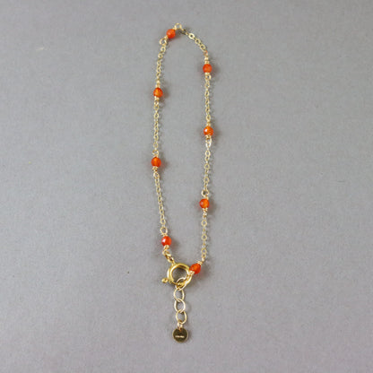 A delicate gold chain bracelet with small orange beads spaced along the chain and a gold clasp with an extender and small hanging tag, shown on a plain gray background.