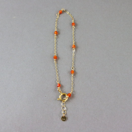 A delicate gold chain bracelet with small orange beads spaced along the chain and a gold clasp with an extender and small hanging tag, shown on a plain gray background.