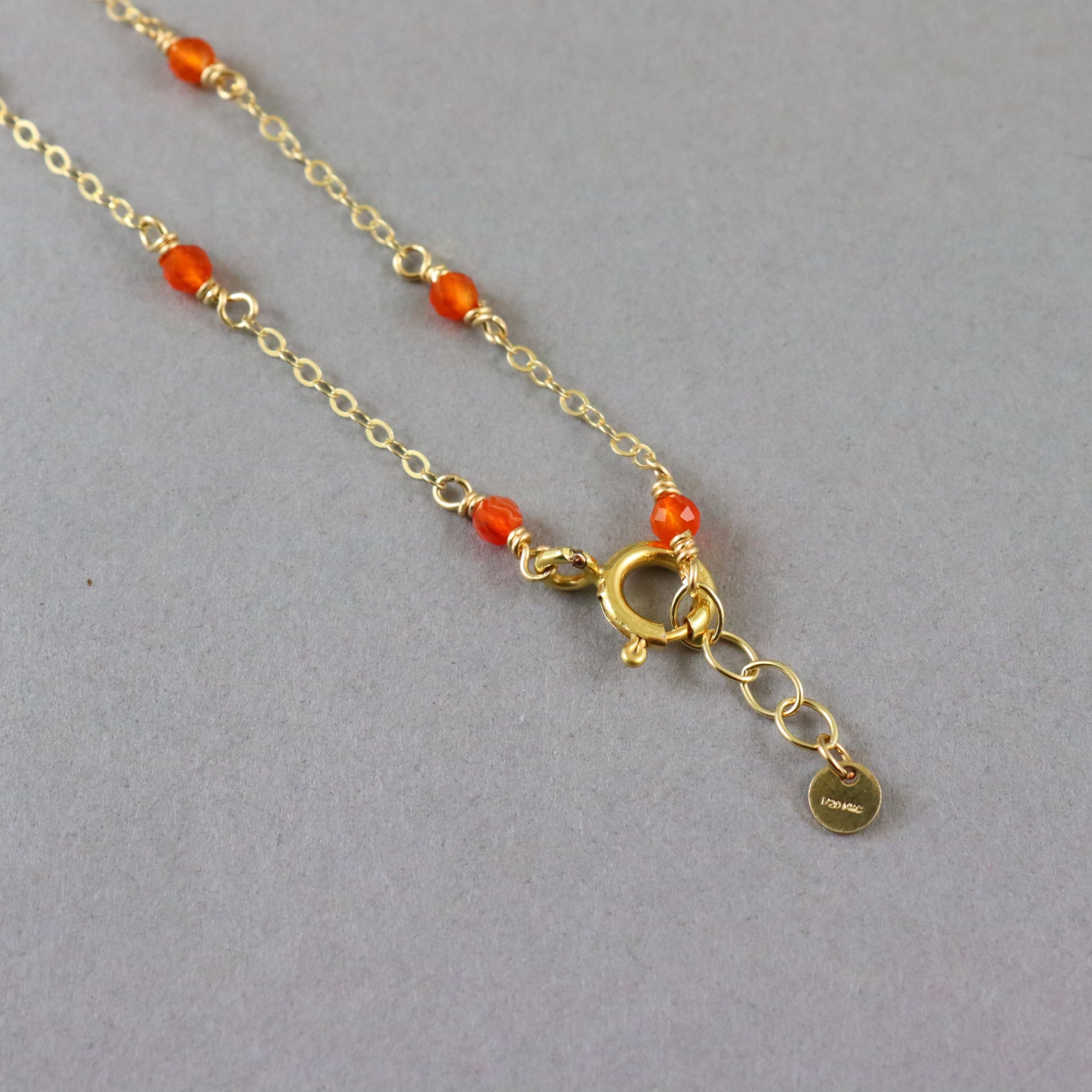 A gold chain necklace with small orange-red beads spaced along the chain, a gold clasp, and an extender chain with a small round tag, photographed on a plain gray background.