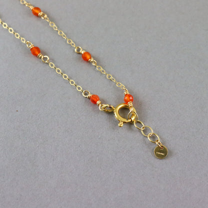 A gold chain necklace with small orange-red beads spaced along the chain, a gold clasp, and an extender chain with a small round tag, photographed on a plain gray background.