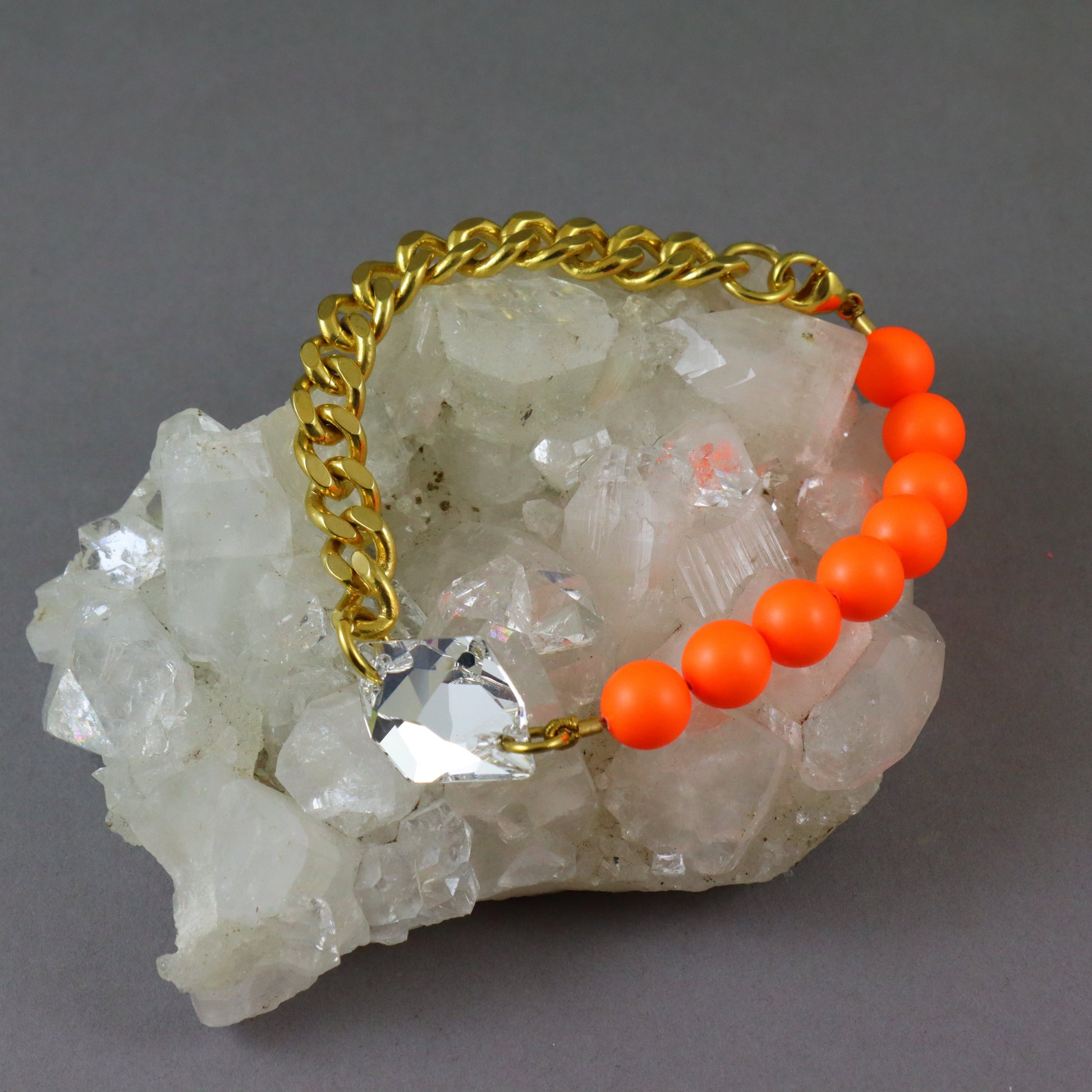 A gold chain bracelet with bright orange round beads and a clear faceted crystal charm, displayed on a white quartz crystal cluster against a gray background.