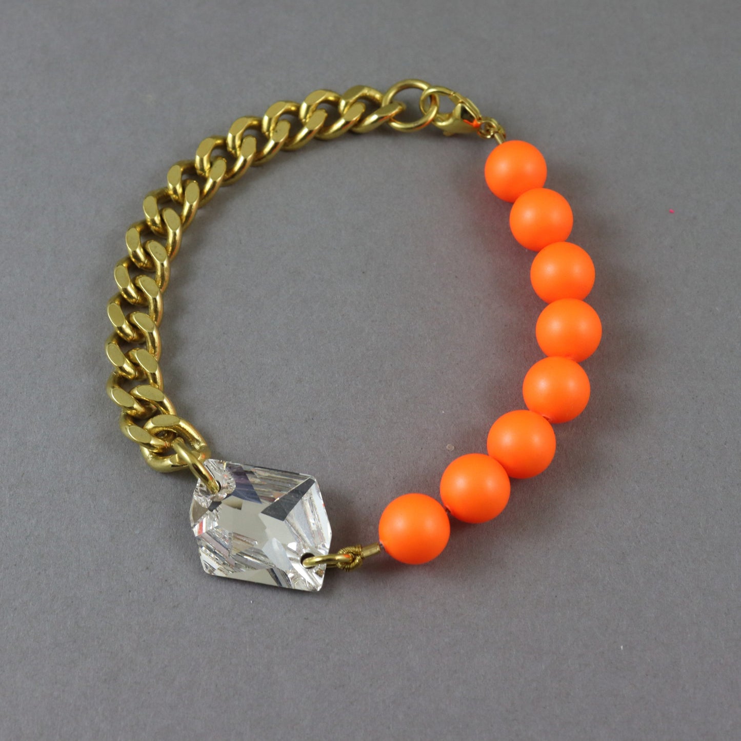 A gold-tone chain bracelet with a row of bright orange round beads and a faceted clear crystal charm, displayed on a neutral gray background.