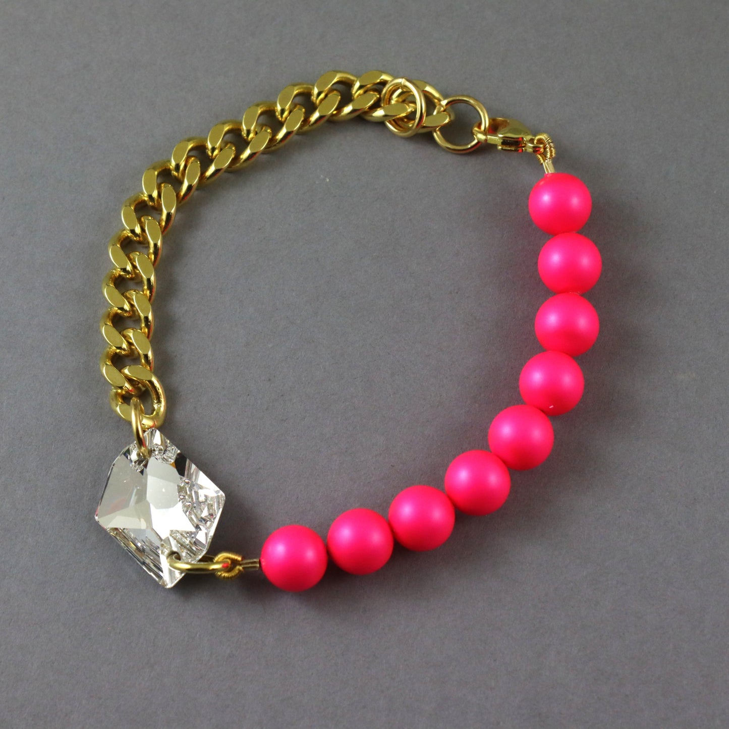A gold-tone chain bracelet featuring a row of bright pink beads and a clear faceted crystal charm, displayed on a neutral gray background.