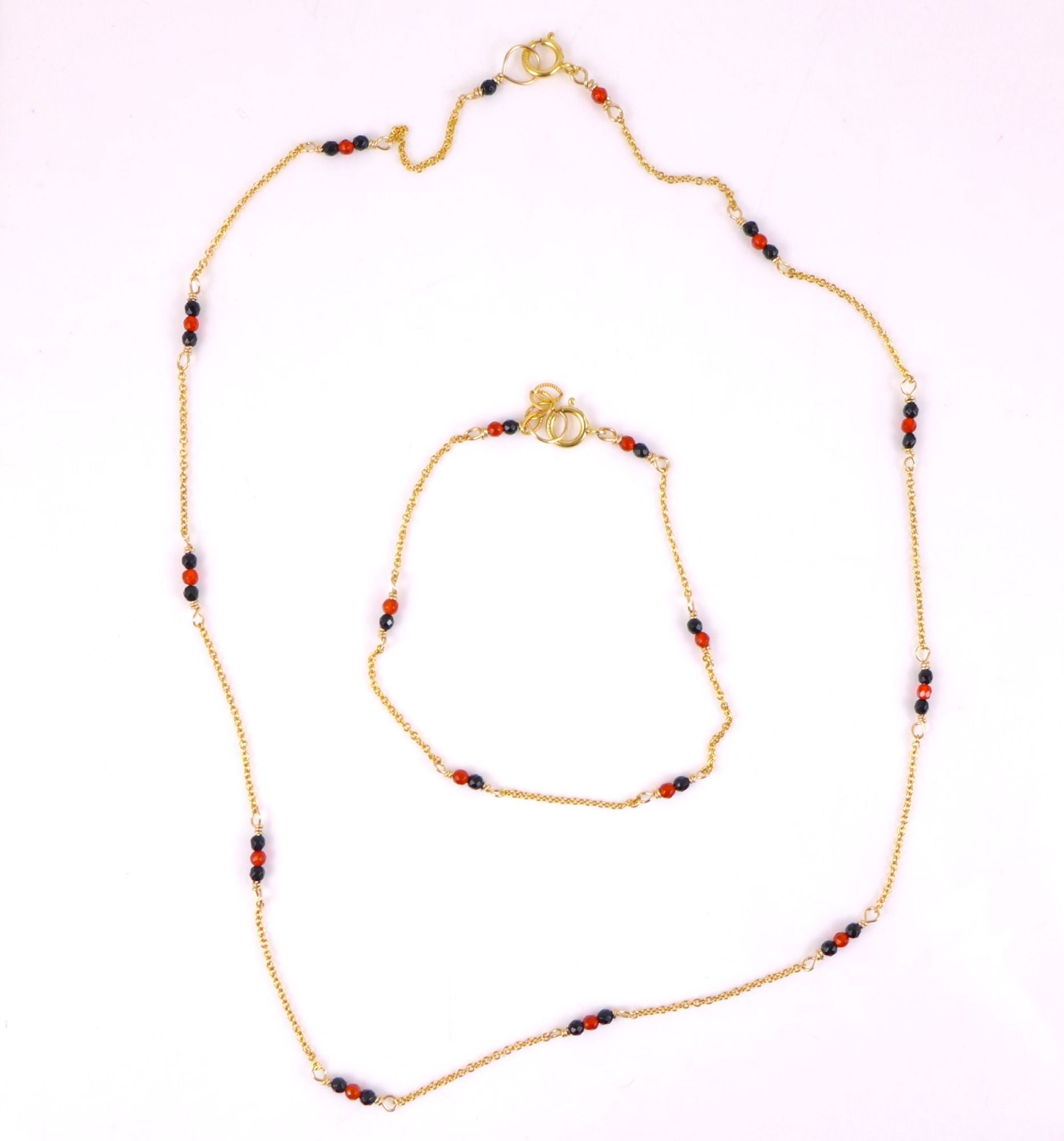 A gold chain necklace and matching bracelet set, each adorned with small beads in red, black, and white, displayed on a light pink background.